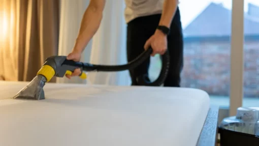 A man vacuuming dirt and dust from a bare mattress in a bedroom