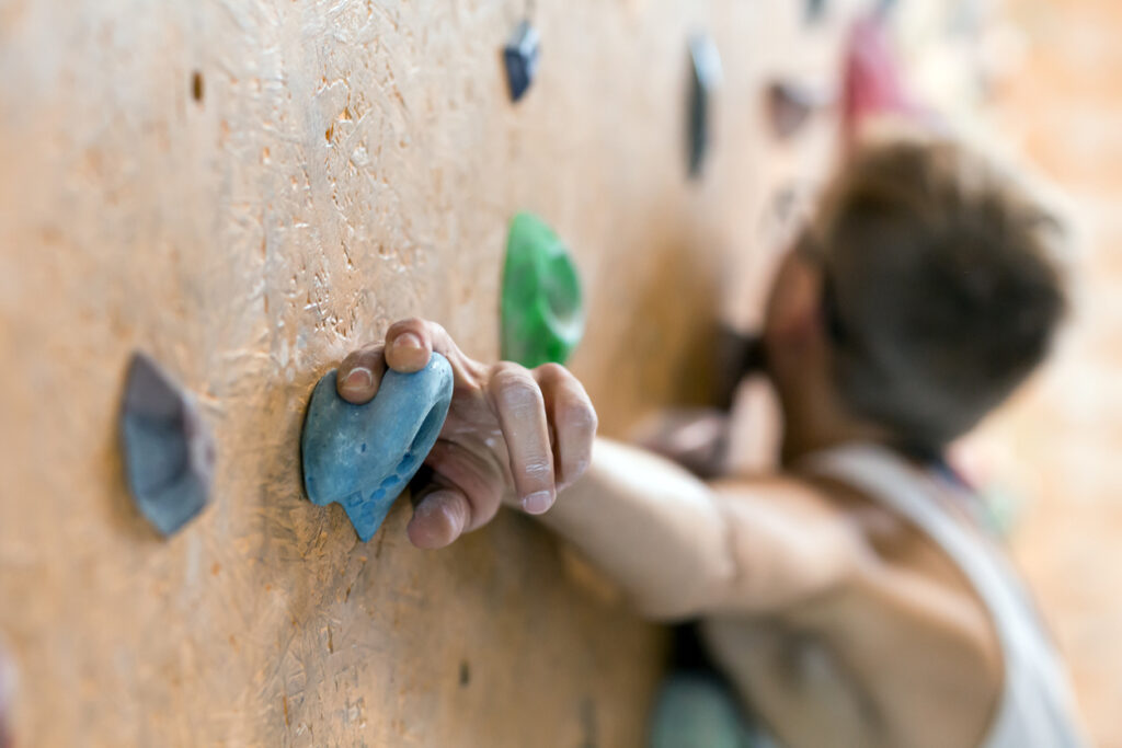 A man in a white tank top climbs up a rock climbing wall. Two fingers on his left hand wrap around a blue rock in front of the camera.