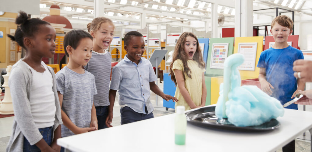 A group of young children gather around a white table during a science demonstration. A girl in a yellow shirt looks surprised as blue foam bubbles out of a beaker.