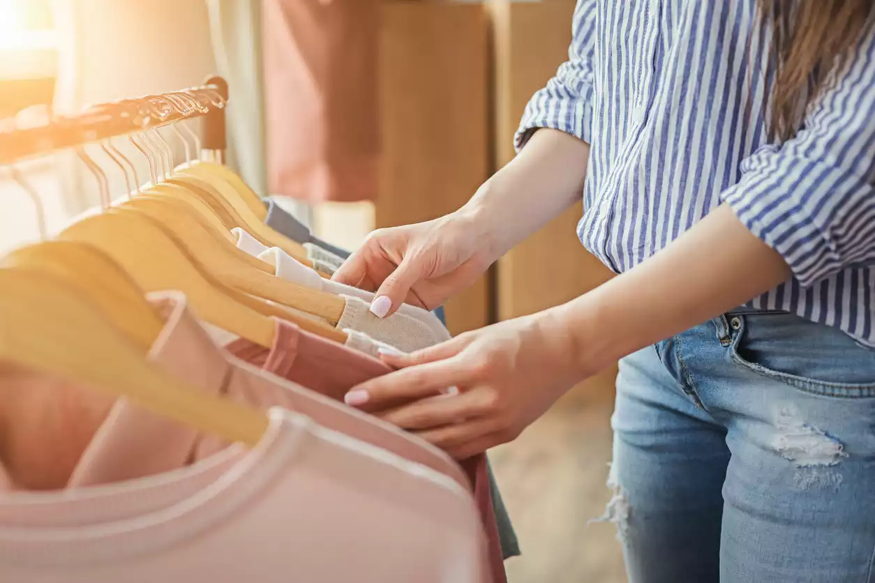 A woman wearing a blue and white striped shirt and denim jeans flips through a rack of pink and nude colored shirts and blouses hanging on a rack in a boutique store.