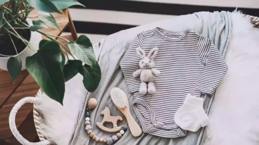 A stuffed rabbit, wooden toys, and brush sit on top of a gray and white striped onesie. These items all lay in a bassinet next to a plant with large, green leaves.