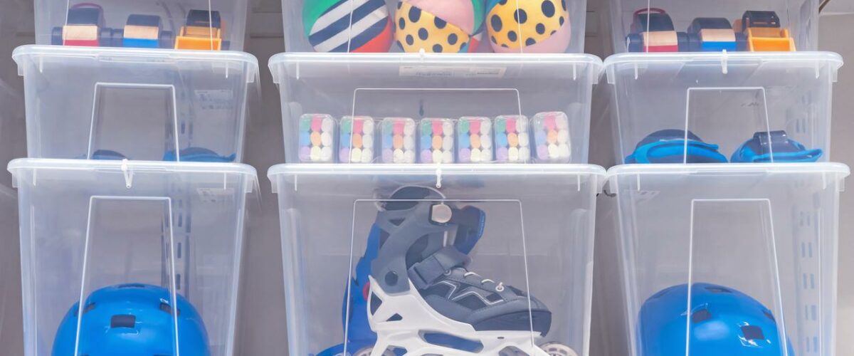 Clear storage bins hold roller skates, helmets, and outdoor toys. The boxes are stacked neatly on a shelf.