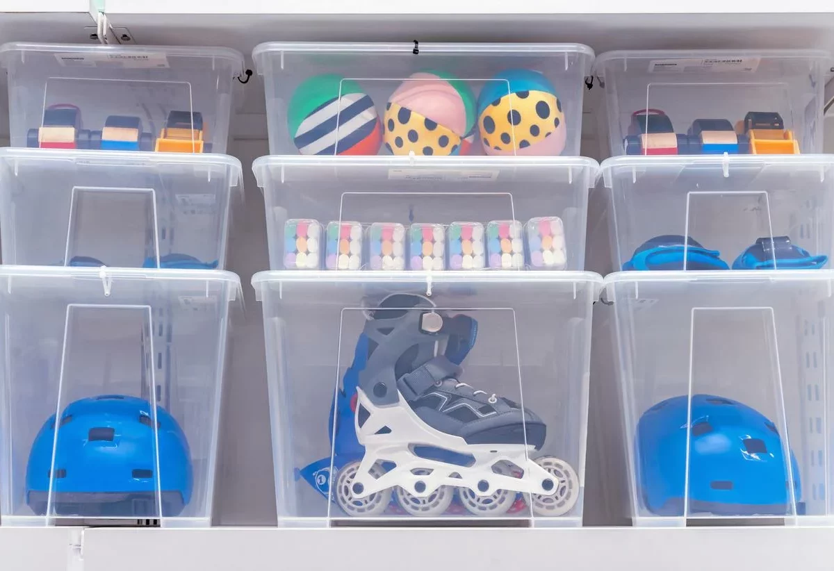 Clear storage bins hold roller skates, helmets, and outdoor toys. The boxes are stacked neatly on a shelf.