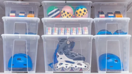 Clear storage bins hold roller skates, helmets, and outdoor toys. The boxes are stacked neatly on a shelf.