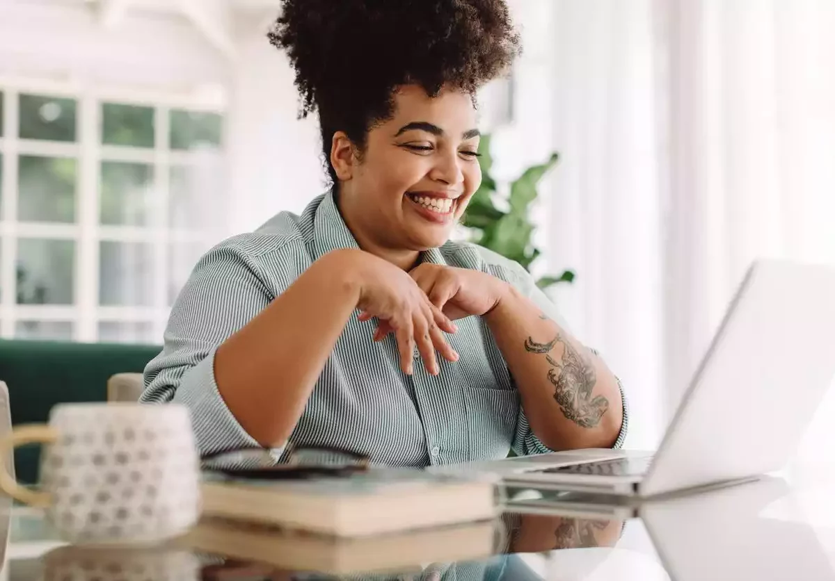 A woman sits at a desk in a brightly lit room and smiles at a laptop in front of her.