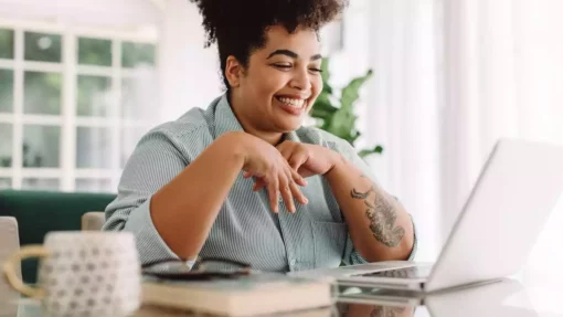 A woman sits at a desk in a brightly lit room and smiles at a laptop in front of her.