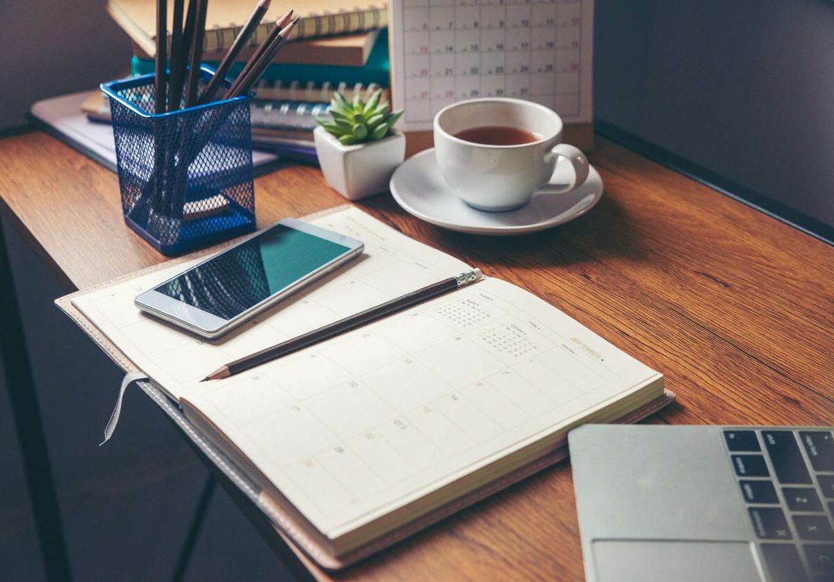 A book sits open on a desk surrounded by a coffee cup, cell phone, laptop, and container of pencils.