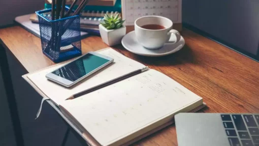 A book sits open on a desk surrounded by a coffee cup, cell phone, laptop, and container of pencils.