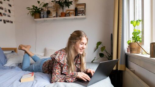 A woman lays on a bed and works on a laptop.