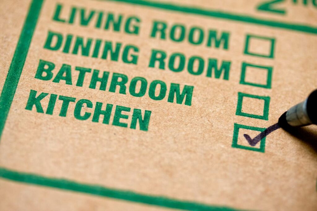 A cardboard box features a list of rooms with the box next to kitchen checked with a black marker