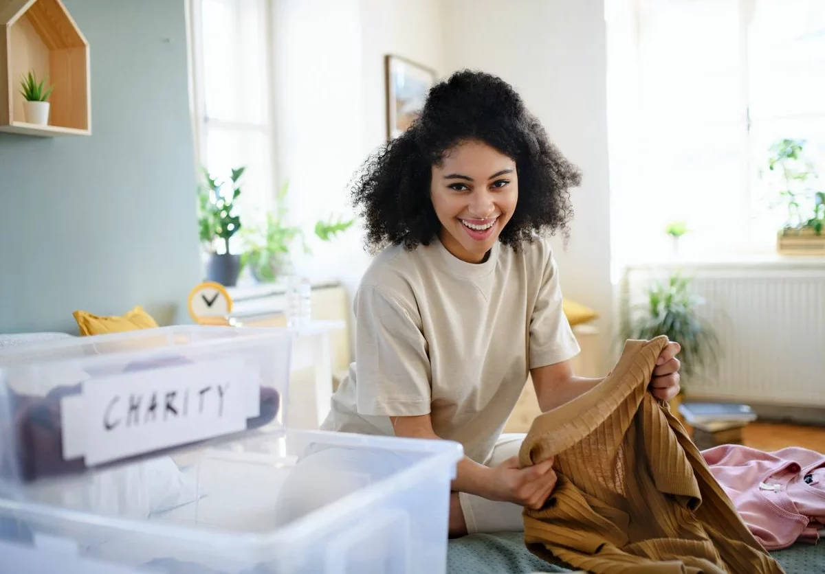 A woman sorts through clothing in a bright room next to a stack of clear storage totes. One tote is labeled “charity”