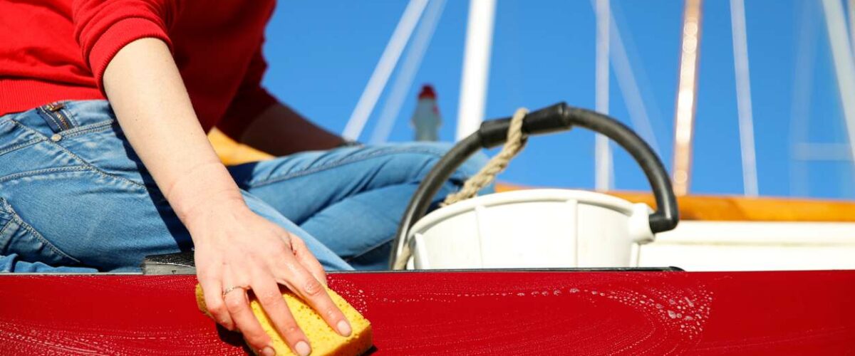 A person sits on the edge of a red boat and cleans it with a yellow sponge and white bucket