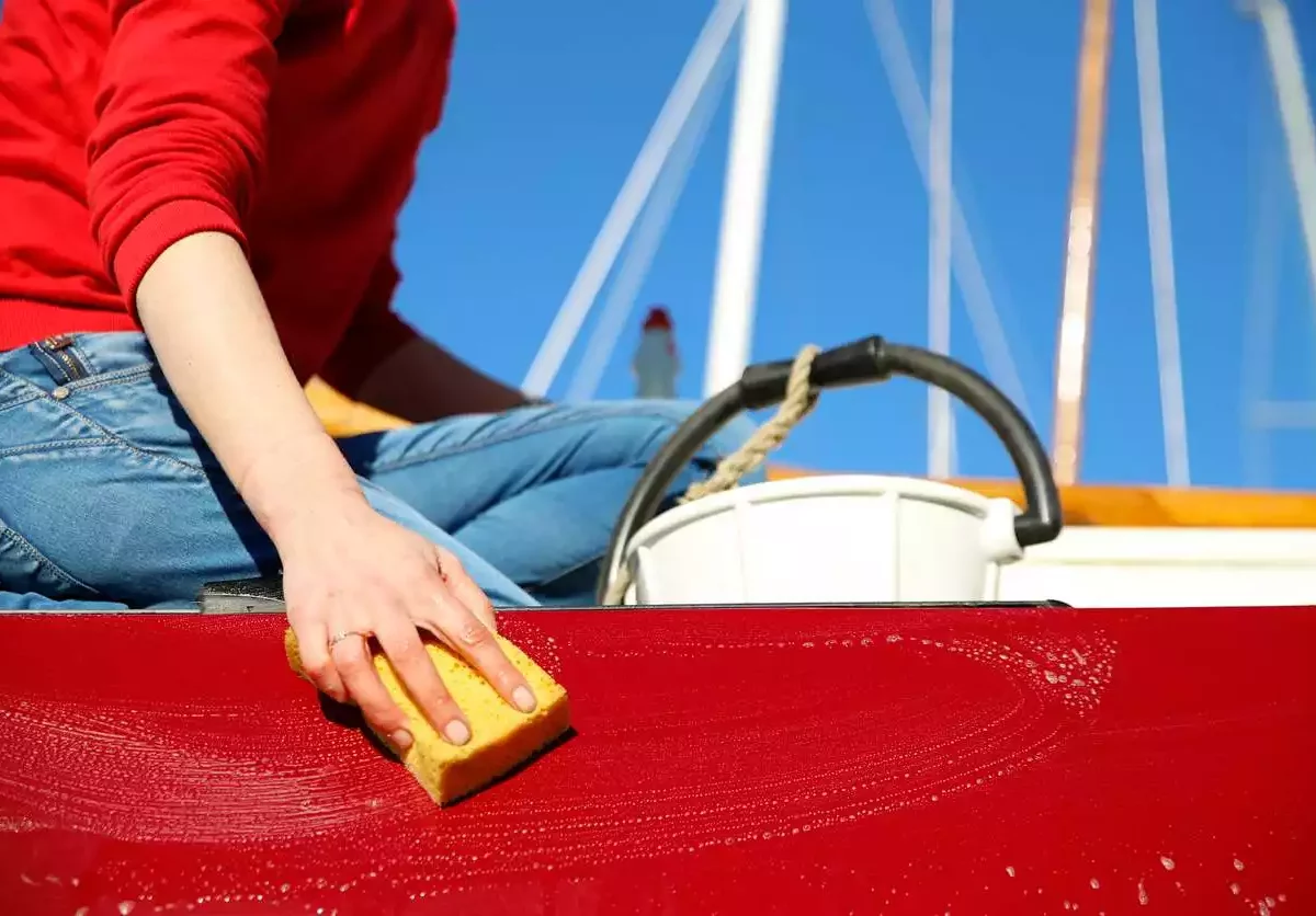 A person sits on the edge of a red boat and cleans it with a yellow sponge and white bucket