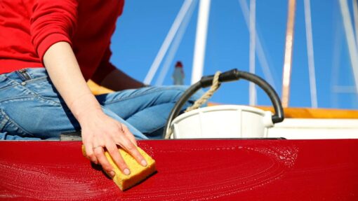 A person sits on the edge of a red boat and cleans it with a yellow sponge and white bucket