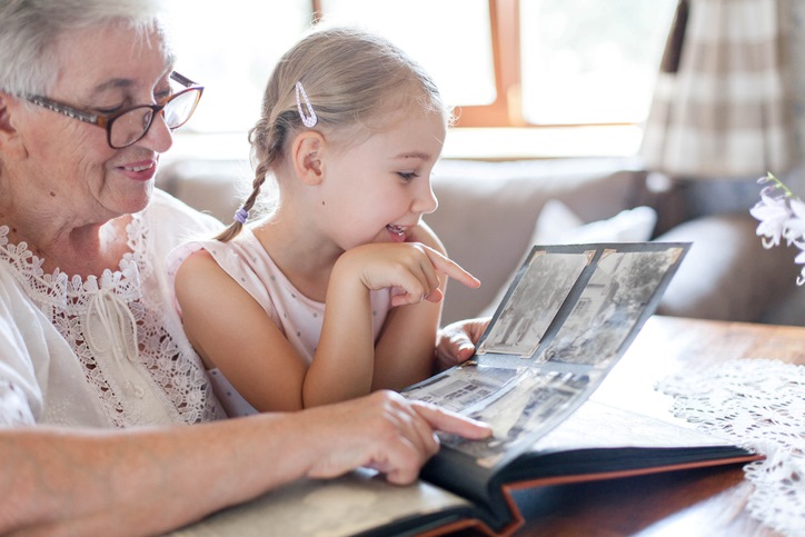An older woman in a white shirt and glasses shows a young girl images in a photo album