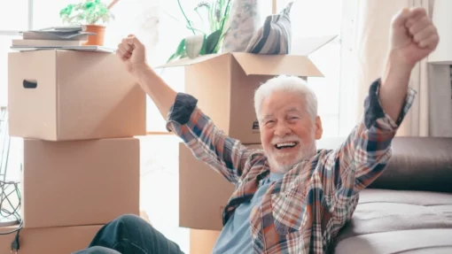 Smiling senior man sitting by the couch in front of cardboard moving boxes.