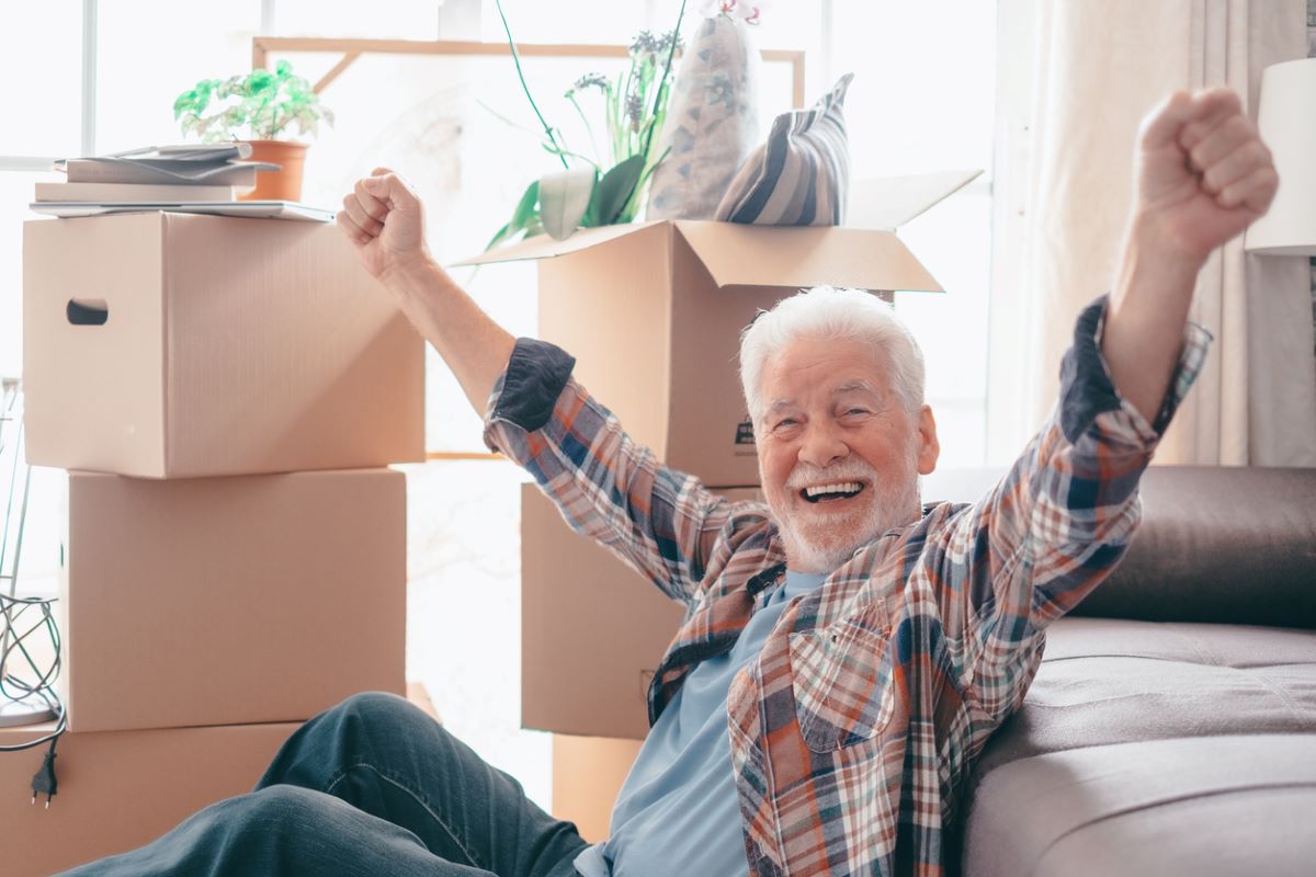 Smiling senior man sitting by the couch in front of cardboard moving boxes.