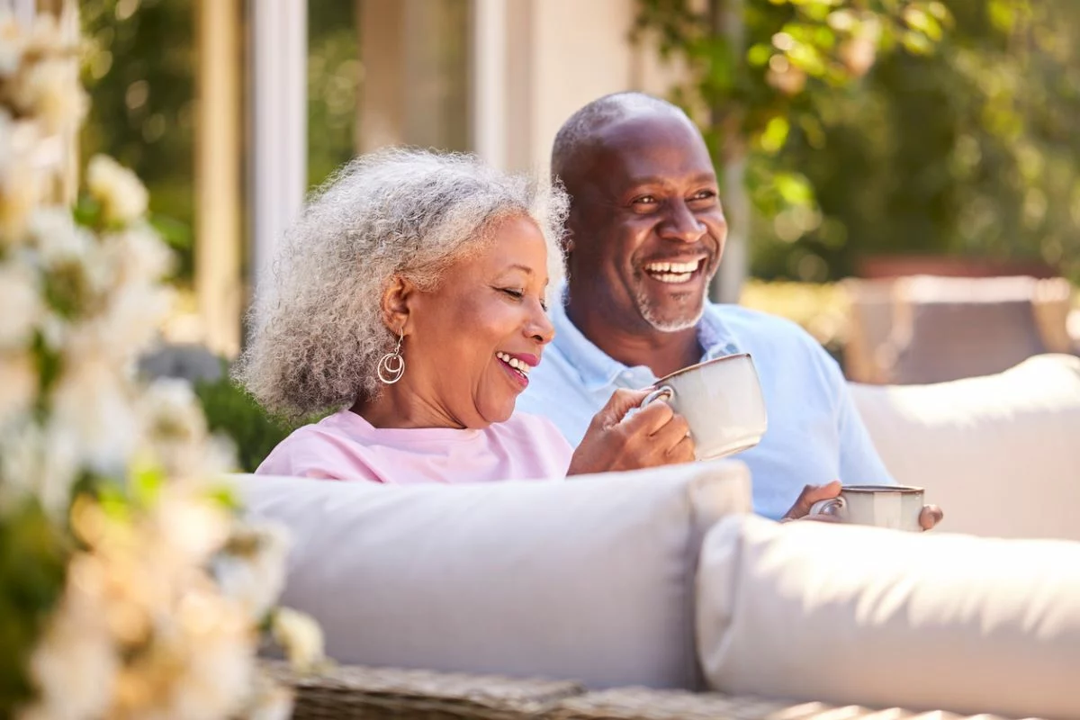 A retired couple sitting outside, smiling, and drinking coffee in the sun.