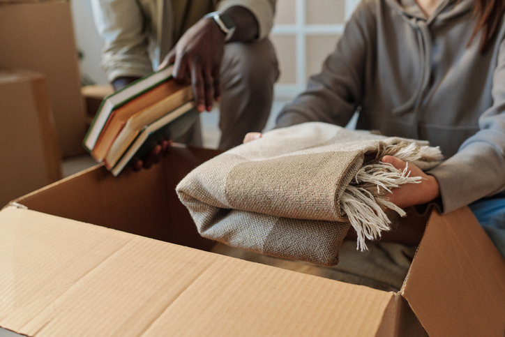 Two people packing belongings, including a blanket and books, into a box for storage.