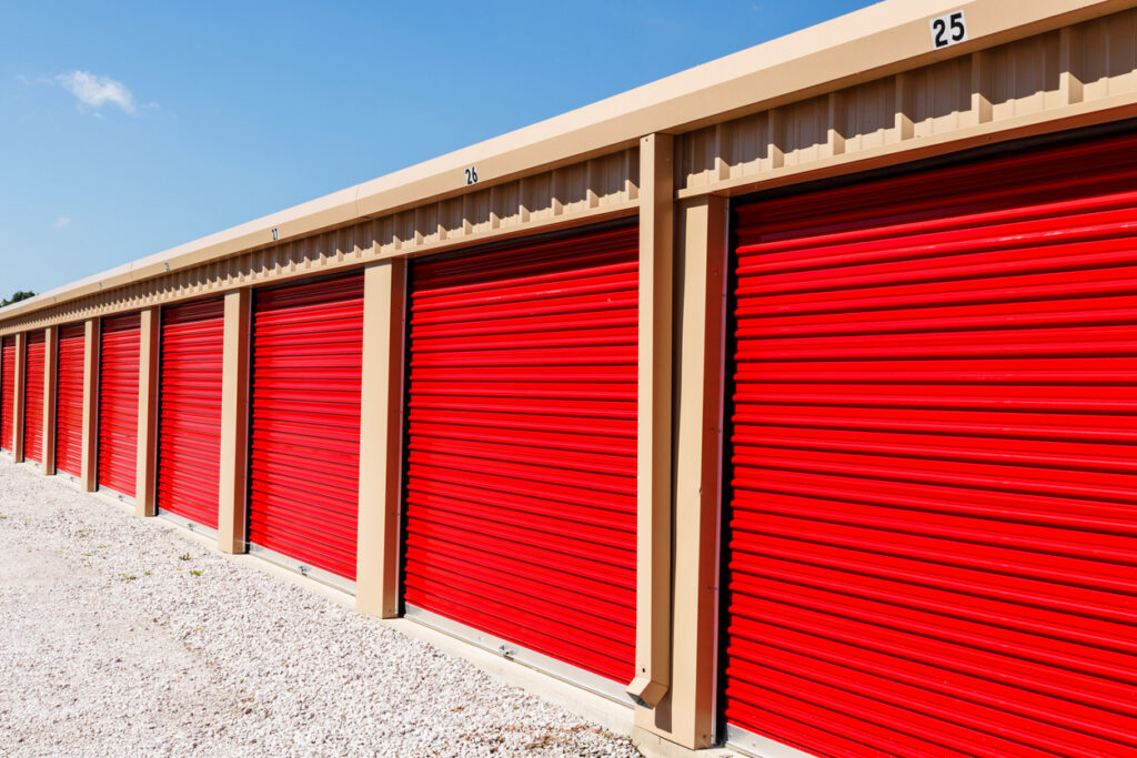 A row of outdoor drive-up storage units with red doors on a clear day.