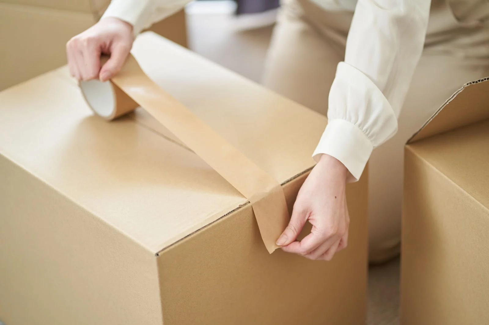 A woman wearing a long-sleeve blouse tapes up a box for storage.