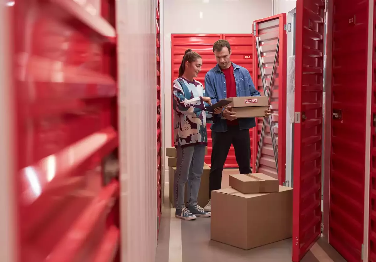 A man and woman load boxes into a storage unit with a red door.