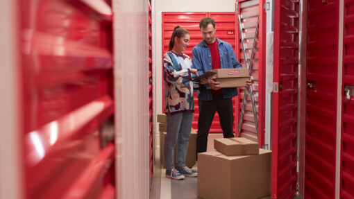 A man and woman load boxes into a storage unit with a red door.
