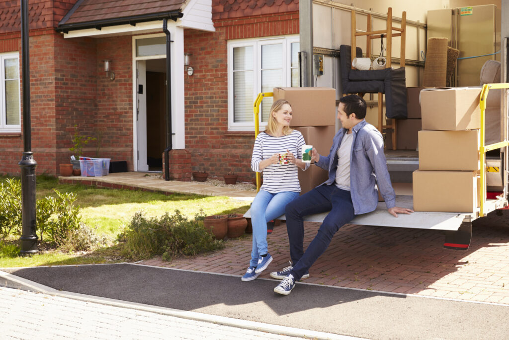 A man and woman sitting outside a moving truck filled with boxes while holding mugs on a sunny day.