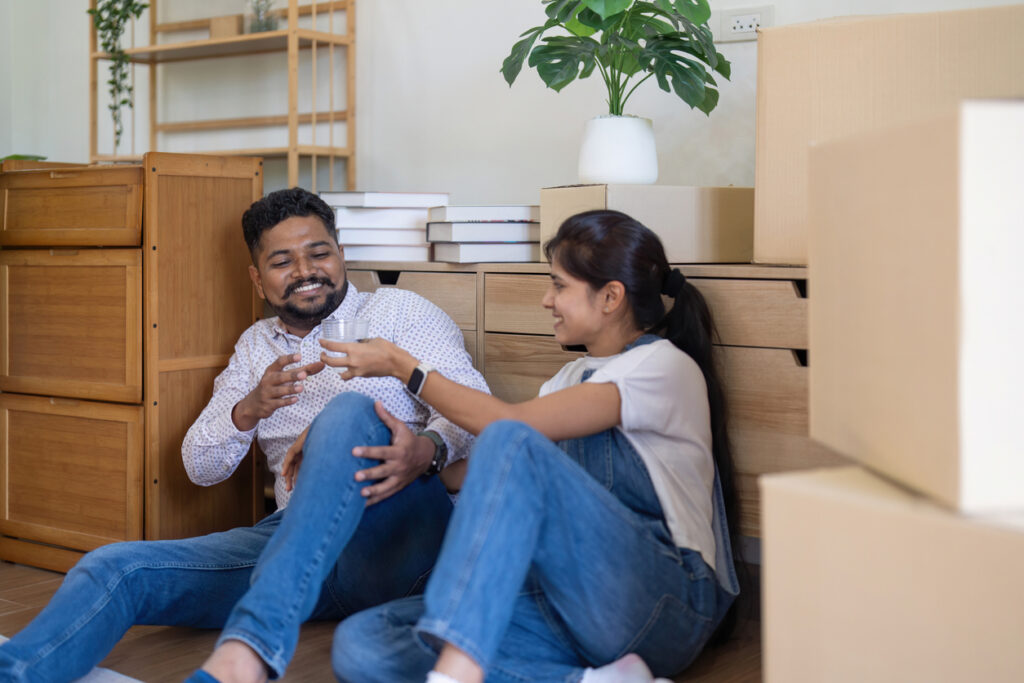 A man and woman drinking water surrounded by furniture and boxes during a move.