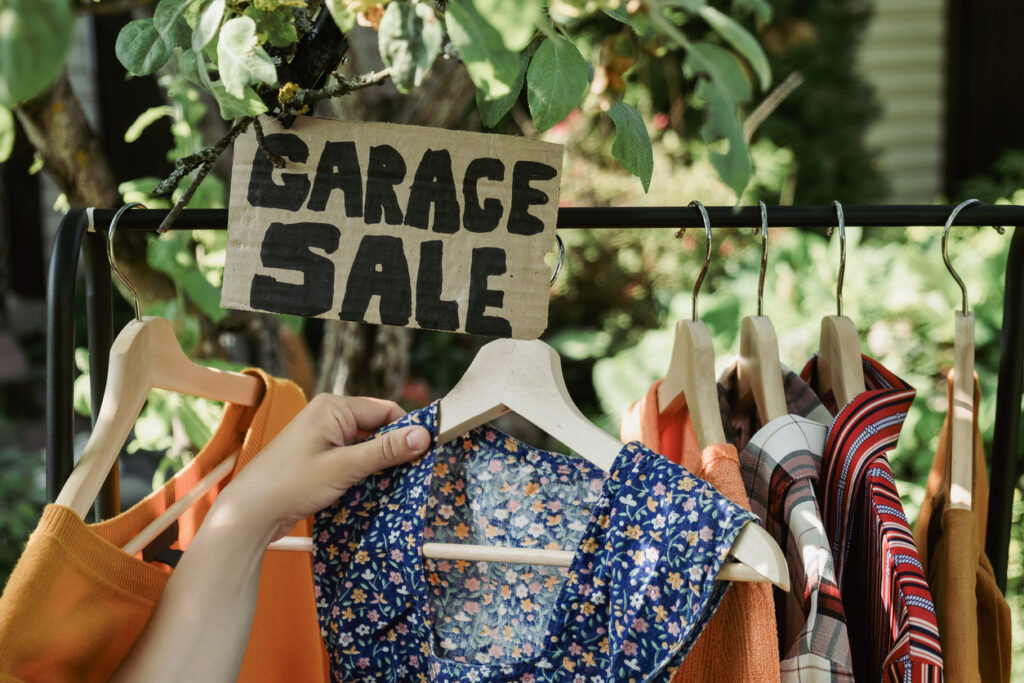 A close-up of hands taking clothes off a clothing rack labeled “Garage Sale” outdoors during the day.