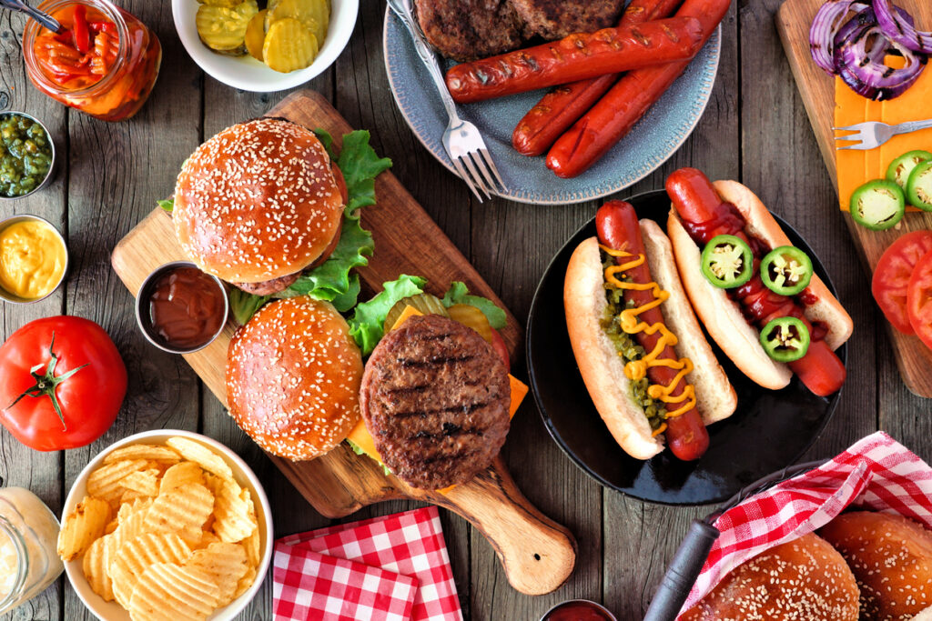 A table of food for a summer barbecue, including hot dogs, burgers, chips, condiments, and toppings.