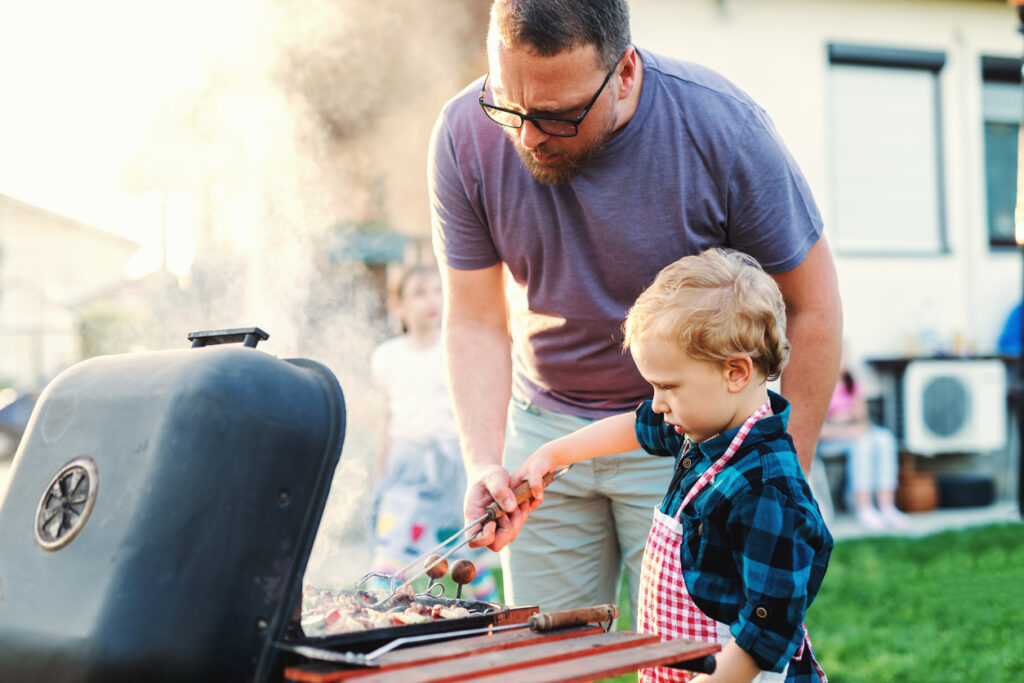 A father teaching his young son to use a grill in the backyard.