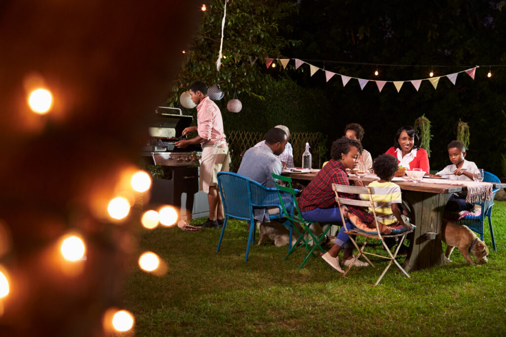 A family enjoying dinner while a father grills in their backyard at night, surrounded by lights and decorations.