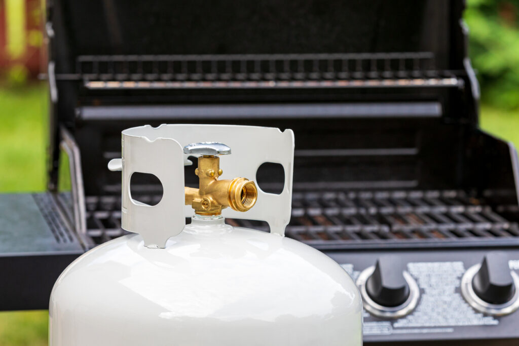 A close-up of a propane gas cylinder in front of a backyard grill.