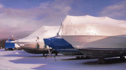 Covered motorboats with snow on top sitting in a snowy parking lot.