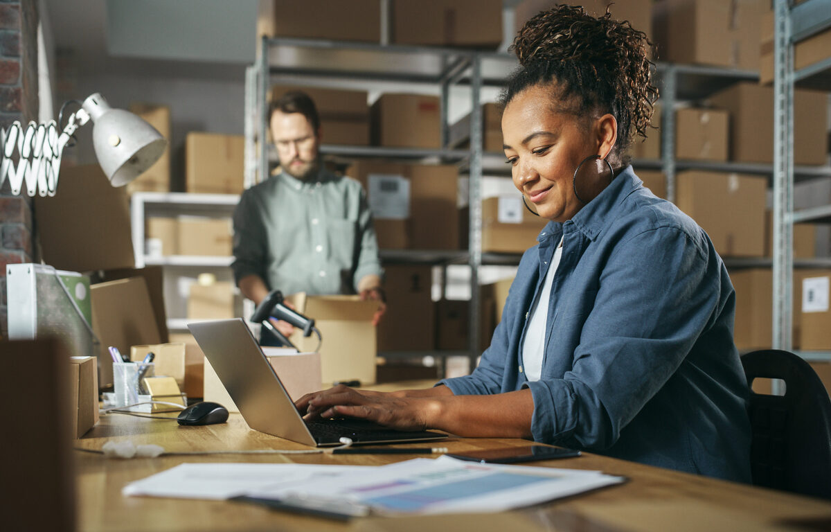 A smiling female business owner typing on her computer with shelves of boxes behind her.