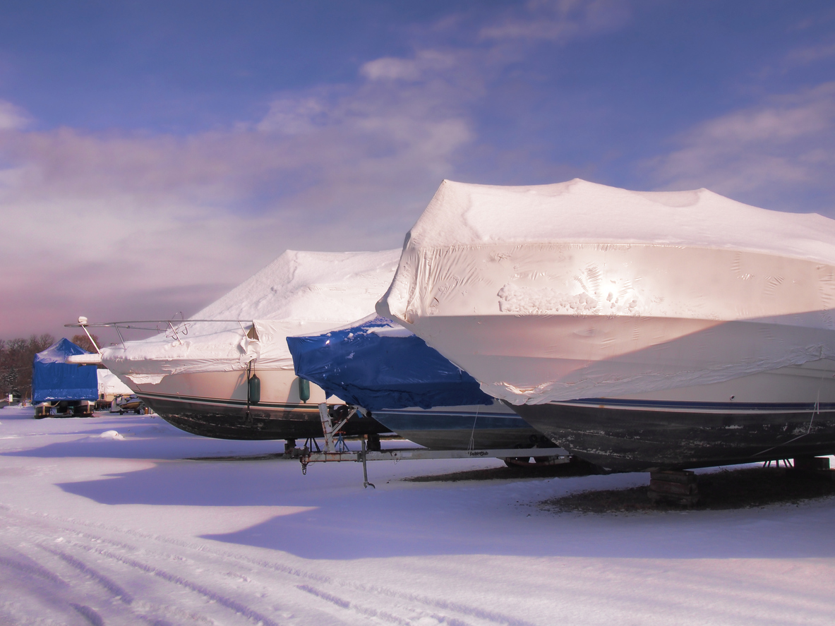 Covered motorboats with snow on top sitting in a snowy parking lot.