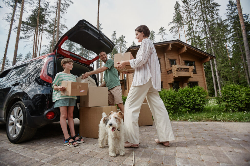 A family loads their car with moving boxes with their dog beside them.