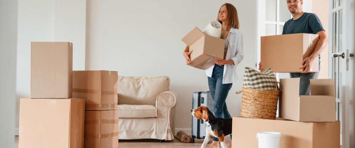 A man and woman holding boxes walk into their new home with their dog.