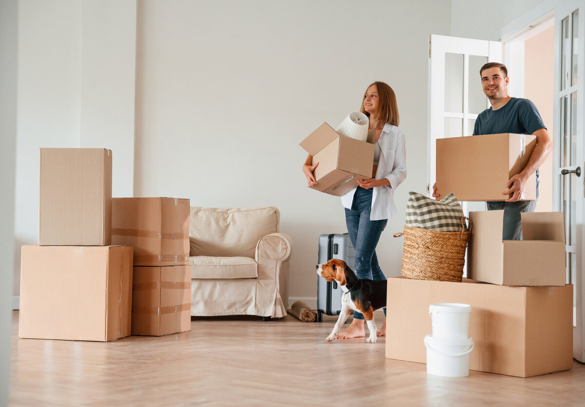 A man and woman holding boxes walk into their new home with their dog.