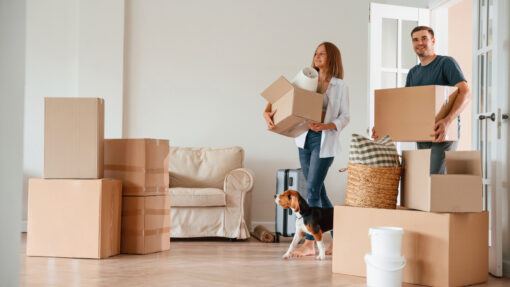 A man and woman holding boxes walk into their new home with their dog.