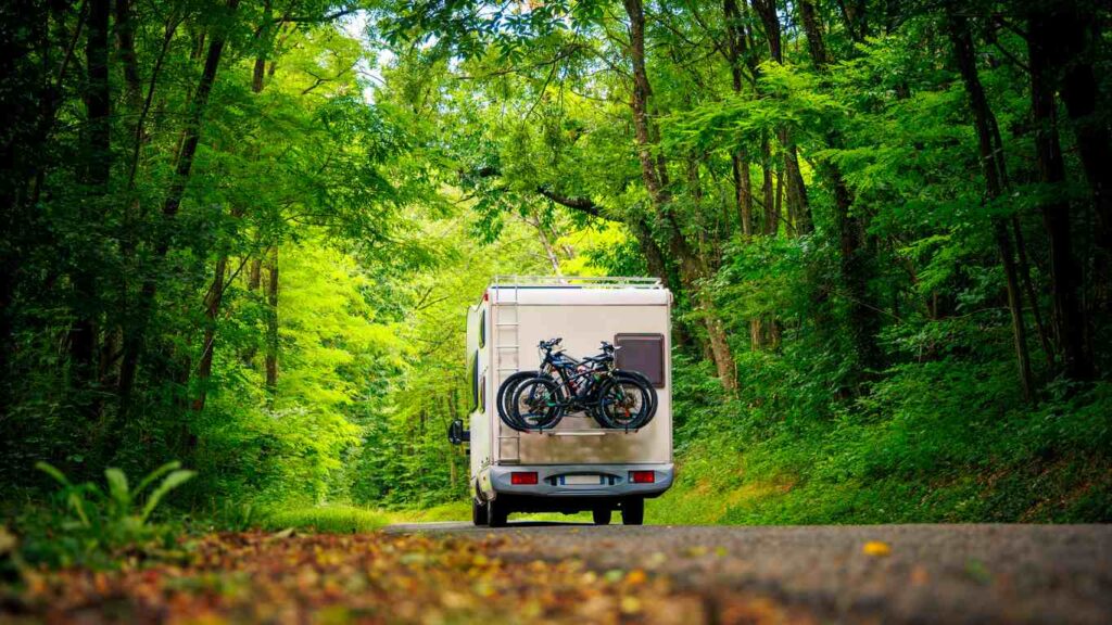 An RV drives through a wooded area with bikes on the back.