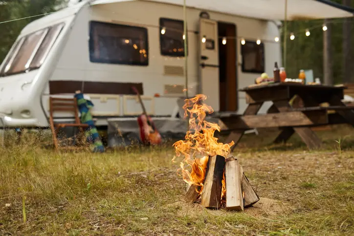 A campfire in front of a white RV with a picnic table set up in a cozy forest campsite.