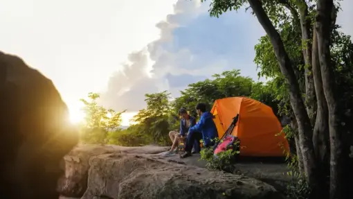 Two campers sit on a rocky cliff near an orange tent at sunrise, surrounded by trees.