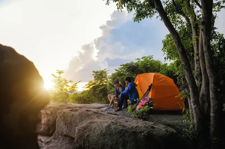Two campers sit on a rocky cliff near an orange tent at sunrise, surrounded by trees.