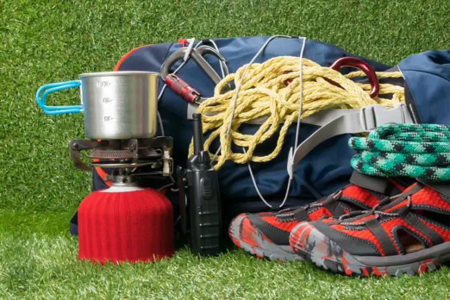 Camping and hiking gear on grass, including a stove, backpack, hiking shoes, and more.