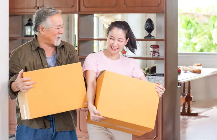 A father and daughter smile together while carrying boxes into their home.