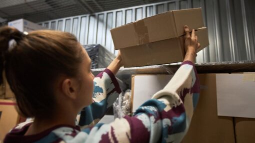 A woman in a sweater places two flat cardboard boxes on a shelf inside a storage unit with boxes stacked nearby.