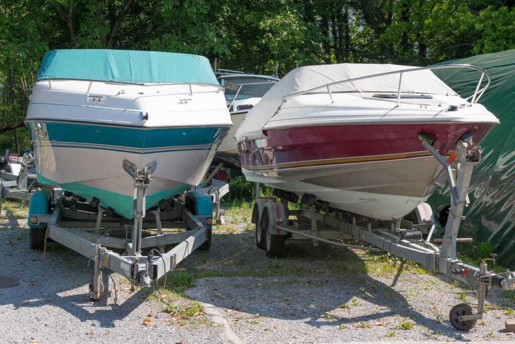 Two boats on trailers with fitted covers, parked outdoors in a storage area surrounded by trees and gravel ground.