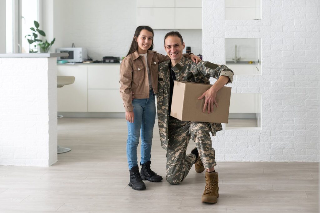 A military member smiles with his daughter while holding a box as they move into their new home.